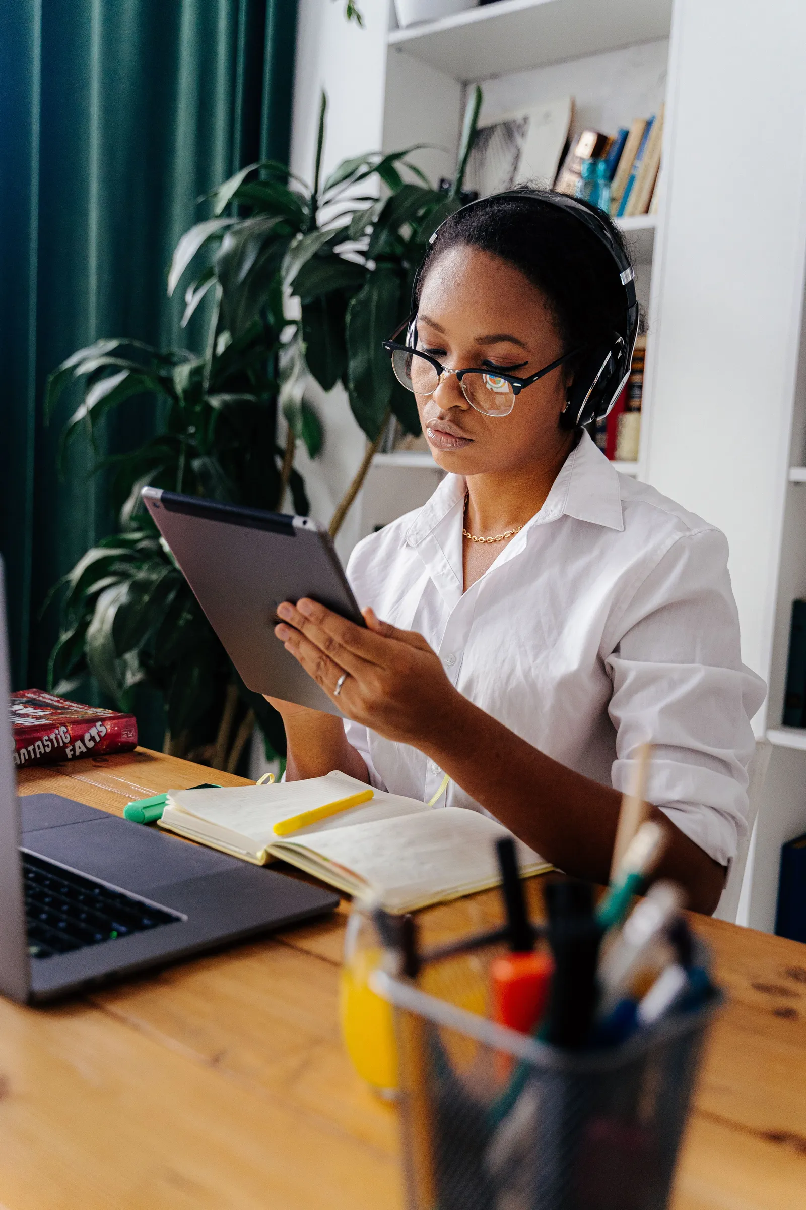 A woman in headphones reading from a tablet at a desk with her laptop and notes open